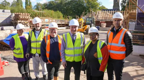 Six people stand in a line, wearing a mixture of yellow and orange hi-vis waistcoats. They are all wearing a white hard hat and standing on a housing development that is work in progress. In the background you can see wooden rafters of roofs being build, with builders and equipment dotted about.